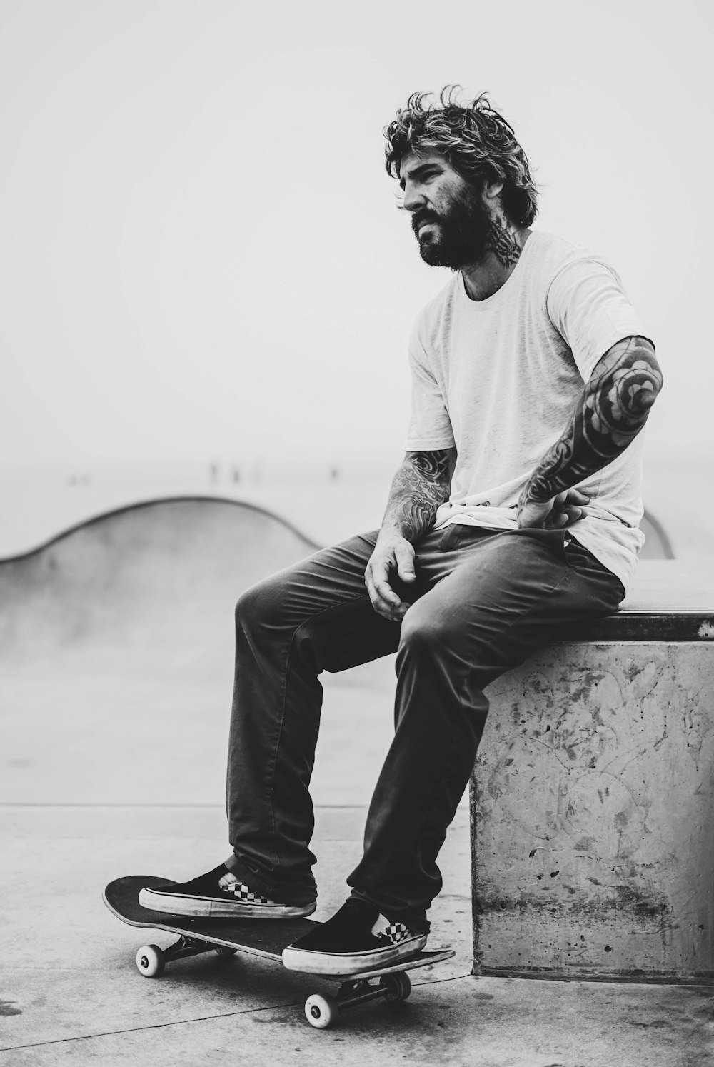 Photographie en niveaux de gris d’un homme assis sur un banc tandis que ses pieds sur une planche à roulettes