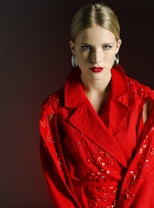 A woman with blonde hair pulled back is wearing a vibrant red coat adorned with intricate embroidery and embellishments. Her bold red lipstick matches the coat, and she accessorizes with large silver earrings. The background is a dark, muted color that enhances the vividness of her attire.