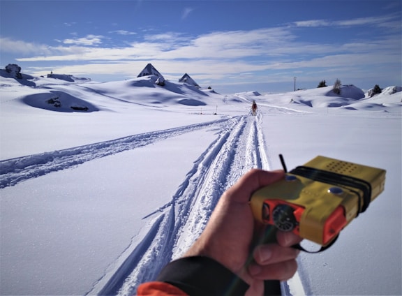 A snow-covered landscape under a clear blue sky with traces of ski tracks leading into the distance. A hand in the foreground holds a yellow device, possibly a camera. In the background, a person skiing away from the camera and snow-covered hills and structures can be seen.
