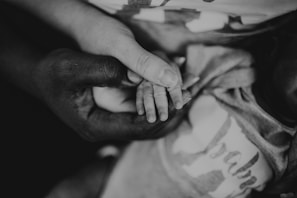 A close-up view of two hands gently holding a child's small hand, creating a sense of care and protection. The image is black and white, adding depth and emotion to the scene.