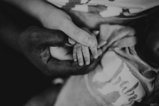 A close-up view of two hands gently holding a child's small hand, creating a sense of care and protection. The image is black and white, adding depth and emotion to the scene.