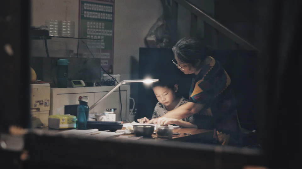 The image shows a woman and a child sitting at a table, both focused on their work. The woman is holding a pen and paper, while the child has a cup of coffee next to them. A lamp stands on the table in front of them, providing light for their task. In the background, there are several books scattered around, suggesting that they might be working on a project or studying together.