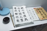 A workspace featuring traditional Chinese calligraphy set on a desk. Sheets of paper with elegant Chinese characters in black ink are arranged alongside a bamboo calligraphy brush and inkstone. A stack of books or practice sheets is placed to the side, and a small blue desk lamp is positioned at the corner.