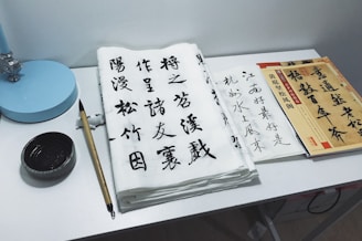 A workspace featuring traditional Chinese calligraphy set on a desk. Sheets of paper with elegant Chinese characters in black ink are arranged alongside a bamboo calligraphy brush and inkstone. A stack of books or practice sheets is placed to the side, and a small blue desk lamp is positioned at the corner.