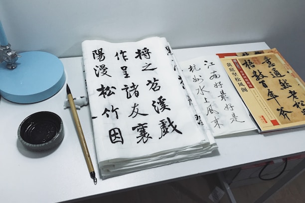 A workspace featuring traditional Chinese calligraphy set on a desk. Sheets of paper with elegant Chinese characters in black ink are arranged alongside a bamboo calligraphy brush and inkstone. A stack of books or practice sheets is placed to the side, and a small blue desk lamp is positioned at the corner.