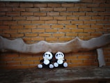 Two plush panda toys with bows around their necks are seated on a rustic, carved wooden bench. The background is a brick wall with a warm, muted tone, complementing the natural wood and soft, plush textures of the toys.