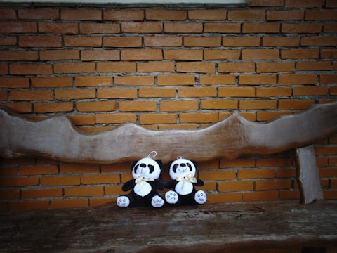 Two plush panda toys with bows around their necks are seated on a rustic, carved wooden bench. The background is a brick wall with a warm, muted tone, complementing the natural wood and soft, plush textures of the toys.