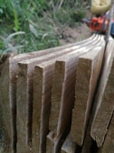 Several raw wooden planks are stacked closely together with visible grain patterns, set against a natural outdoor background. A blurred chainsaw is partially visible in the top right corner, adding to the rustic and manual labor atmosphere.