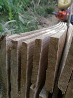 Several raw wooden planks are stacked closely together with visible grain patterns, set against a natural outdoor background. A blurred chainsaw is partially visible in the top right corner, adding to the rustic and manual labor atmosphere.