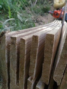 Several raw wooden planks are stacked closely together with visible grain patterns, set against a natural outdoor background. A blurred chainsaw is partially visible in the top right corner, adding to the rustic and manual labor atmosphere.