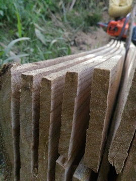 Several raw wooden planks are stacked closely together with visible grain patterns, set against a natural outdoor background. A blurred chainsaw is partially visible in the top right corner, adding to the rustic and manual labor atmosphere.