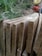 Several raw wooden planks are stacked closely together with visible grain patterns, set against a natural outdoor background. A blurred chainsaw is partially visible in the top right corner, adding to the rustic and manual labor atmosphere.