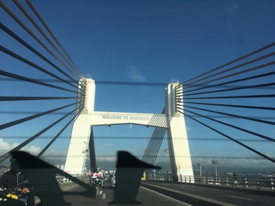 A bridge with prominent cable wires stretches across a vast expanse under a clear blue sky. The structure features large white pillars with signage that reads 'WELCOME TO MANDAUE'. Motorcyclists and vehicles can be seen traveling across the bridge.