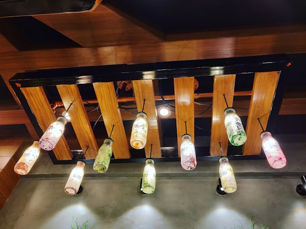 A ceiling light fixture made from wooden slats, featuring hanging bottles that serve as light covers. The bottles are decorated with colorful paint splatters in shades of red, green, and yellow, and are suspended by black cords.