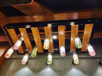 A ceiling light fixture made from wooden slats, featuring hanging bottles that serve as light covers. The bottles are decorated with colorful paint splatters in shades of red, green, and yellow, and are suspended by black cords.