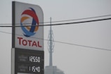 A tall sign for Total with the company's logo and current petrol and diesel prices. The backdrop includes an overcast sky and visible power lines overhead.