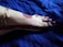 A foot is adorned with intricate henna designs, featuring a bold strip running along the ankle and extending to the toes. A delicate gold anklet with blue and green gemstones enhances the aesthetic, set against a deep blue fabric background.