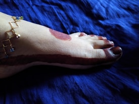 A foot is adorned with intricate henna designs, featuring a bold strip running along the ankle and extending to the toes. A delicate gold anklet with blue and green gemstones enhances the aesthetic, set against a deep blue fabric background.