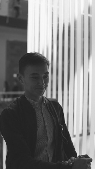 A person wearing a jacket over a striped shirt is positioned near vertical light strips, creating a pattern of shadows across their face. The atmosphere is modern and artistic, with a blurred background adding depth.