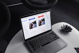 A laptop on a round, white table displaying a design or layout featuring two profiles on the screen. The surrounding environment includes a dark, modern chair and a decorative black item resembling a lotus flower on the table.