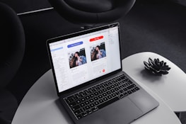 A laptop on a round, white table displaying a design or layout featuring two profiles on the screen. The surrounding environment includes a dark, modern chair and a decorative black item resembling a lotus flower on the table.
