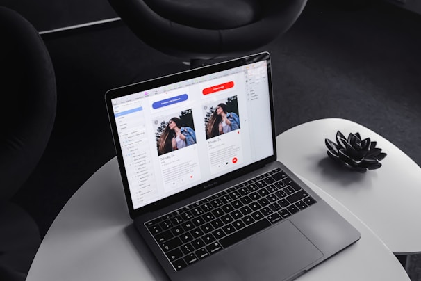 A laptop on a round, white table displaying a design or layout featuring two profiles on the screen. The surrounding environment includes a dark, modern chair and a decorative black item resembling a lotus flower on the table.