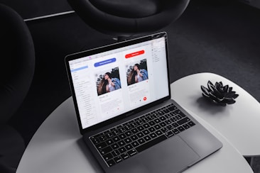 A laptop on a round, white table displaying a design or layout featuring two profiles on the screen. The surrounding environment includes a dark, modern chair and a decorative black item resembling a lotus flower on the table.