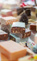 A collection of artisanal soap bars with various patterns, including swirls and geometric prints, stacked and displayed. The focus is on the texture and diversity of the designs, suggesting a handcrafted, festive theme with a pine cone in the background and colorful decorative items scattered around.