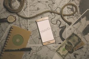 A flat lay composition featuring a smartphone with the word 'TRAVEL' displayed on its screen, positioned on top of several maps. A coiled piece of rope lies across the scene, accompanied by a wristwatch, a notebook with a pen, and some currency including paper bills and coins. The overall theme suggests preparation or planning for a journey.