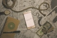 A flat lay composition featuring a smartphone with the word 'TRAVEL' displayed on its screen, positioned on top of several maps. A coiled piece of rope lies across the scene, accompanied by a wristwatch, a notebook with a pen, and some currency including paper bills and coins. The overall theme suggests preparation or planning for a journey.