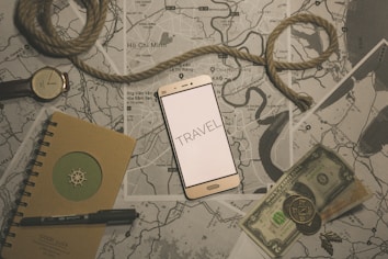 A flat lay composition featuring a smartphone with the word 'TRAVEL' displayed on its screen, positioned on top of several maps. A coiled piece of rope lies across the scene, accompanied by a wristwatch, a notebook with a pen, and some currency including paper bills and coins. The overall theme suggests preparation or planning for a journey.