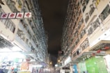 Two tall residential buildings with numerous air conditioning units line a narrow street at night. The lower part of the buildings hosts shops with bright lights, and a few people are visible, some shopping at the stalls. The sky is dark, suggesting it is nighttime.