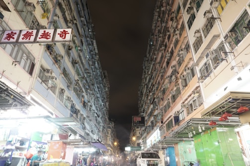 Two tall residential buildings with numerous air conditioning units line a narrow street at night. The lower part of the buildings hosts shops with bright lights, and a few people are visible, some shopping at the stalls. The sky is dark, suggesting it is nighttime.