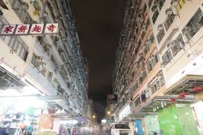 Two tall residential buildings with numerous air conditioning units line a narrow street at night. The lower part of the buildings hosts shops with bright lights, and a few people are visible, some shopping at the stalls. The sky is dark, suggesting it is nighttime.