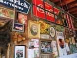 A rustic wall adorned with vintage signs, framed photographs, old advertisements and posters. Notable elements include a large Coca-Cola sign, an image of a historical figure in a portrait, animal pelt, various signs related to batteries and tires, and several promotional materials for an orchard. The wall suggests a nostalgic atmosphere with a mix of personal and promotional memorabilia.