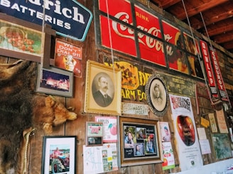 A rustic wall adorned with vintage signs, framed photographs, old advertisements and posters. Notable elements include a large Coca-Cola sign, an image of a historical figure in a portrait, animal pelt, various signs related to batteries and tires, and several promotional materials for an orchard. The wall suggests a nostalgic atmosphere with a mix of personal and promotional memorabilia.