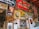 A rustic wall adorned with vintage signs, framed photographs, old advertisements and posters. Notable elements include a large Coca-Cola sign, an image of a historical figure in a portrait, animal pelt, various signs related to batteries and tires, and several promotional materials for an orchard. The wall suggests a nostalgic atmosphere with a mix of personal and promotional memorabilia.