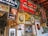 A rustic wall adorned with vintage signs, framed photographs, old advertisements and posters. Notable elements include a large Coca-Cola sign, an image of a historical figure in a portrait, animal pelt, various signs related to batteries and tires, and several promotional materials for an orchard. The wall suggests a nostalgic atmosphere with a mix of personal and promotional memorabilia.