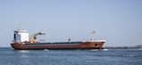 A large cargo ship with an orange hull and white superstructure is sailing through calm blue waters. The sky is clear and the ship is moving towards a breakwater in the background. A small motorboat is passing by the bow of the cargo ship.