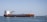 A large cargo ship with an orange hull and white superstructure is sailing through calm blue waters. The sky is clear and the ship is moving towards a breakwater in the background. A small motorboat is passing by the bow of the cargo ship.