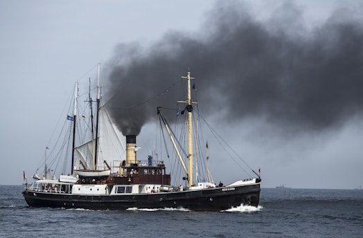 A vintage steamship travels across the ocean, emitting a large plume of black smoke from its chimney. The ship features classic masts and a robust hull, and crew members are visible on deck. The sea is calm, and there is a faint view of the horizon in the background.
