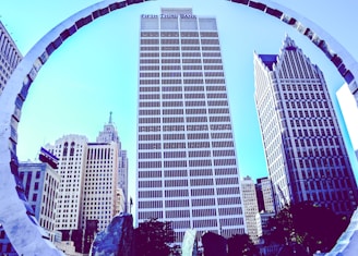 A cityscape featuring tall skyscrapers with distinct architectural designs. A large modern sculpture frames the buildings, creating a unique perspective. The central building displays the 'Fifth Third Bank' logo prominently at the top.