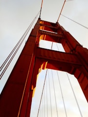 A majestic, towering bridge structure with prominent red-orange pillars and cables reaching towards the sky. The perspective from below accentuates the height and grandeur of the architecture against a light sky background.