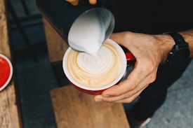 A person is pouring steamed milk from a black pitcher into a cup of coffee, creating latte art in a red cup. The person's hand is visible, wearing a black watch, and the coffee is placed on a wooden surface.