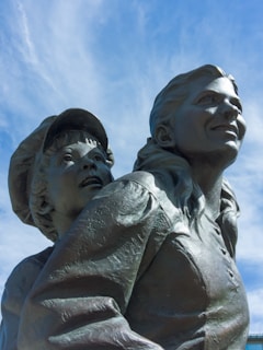 A bronze statue depicting a woman and a child, both looking upward with expressions of hope and determination. The woman's hair is pulled back, and she wears a shirt with visible buttons. The child, wearing a cap, stands slightly behind her, peering over her shoulder.