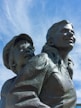 A bronze statue depicting a woman and a child, both looking upward with expressions of hope and determination. The woman's hair is pulled back, and she wears a shirt with visible buttons. The child, wearing a cap, stands slightly behind her, peering over her shoulder.