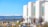 A large, modern building with white stone exterior and tall windows stands on a hillside. The area is landscaped with bushes and trees, and several lampposts line a pathway. In the background, a cityscape is visible, along with a range of snow-capped mountains under a clear blue sky.