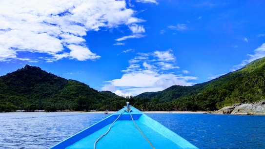 A vibrant blue sky with scattered white clouds stretches over a scenic landscape of lush green hills and a calm body of water. The bow of a boat, painted a bright blue, points towards the shoreline, suggesting a peaceful journey towards the beach. The island is covered with dense greenery and surrounded by a serene, clear blue sea. Rocky outcrops are visible along the edge of the water.