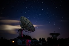 A nighttime scene featuring two large satellite dishes silhouetted against a starry sky. The foreground dish is prominently lit, while a warm glow emanates from a building beneath it. The sky is clear with scattered bright stars, and a few clouds add depth to the composition. Trees line the horizon, adding contrast to the structures.