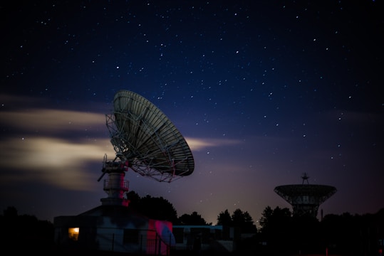 A nighttime scene featuring two large satellite dishes silhouetted against a starry sky. The foreground dish is prominently lit, while a warm glow emanates from a building beneath it. The sky is clear with scattered bright stars, and a few clouds add depth to the composition. Trees line the horizon, adding contrast to the structures.